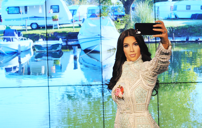Super lifelike Madame Tussauds wax figure of Kim Kardashian superimposed in front of a screen displaying a trailer park